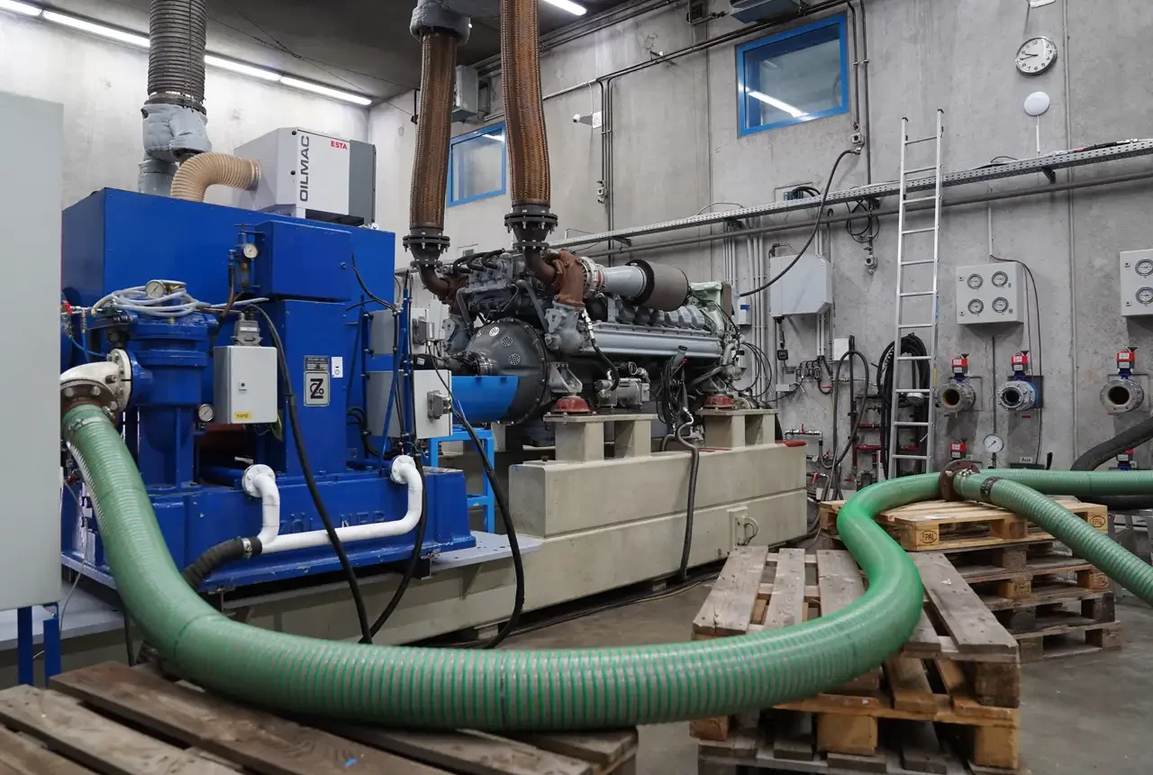 Industrieanlage mit einem großen Motor und Schläuchen in einem Prüfstandraum, umgeben von technischen Geräten und Paletten.