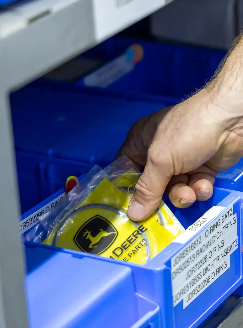 Hand greift nach einem John Deere Ersatzteil in einer blauen Lagerbox.