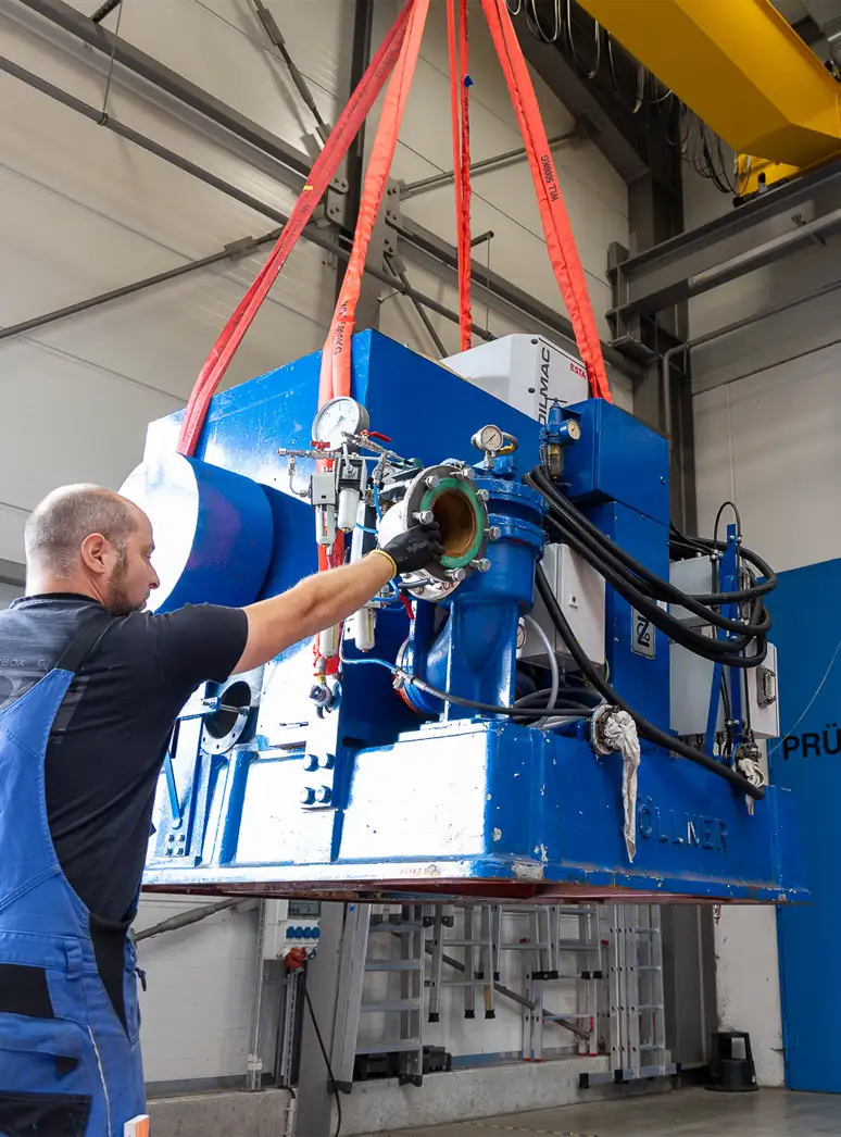Ein Mann in Arbeitskleidung bedient eine große blaue Industriemaschine, die an einem Kran hängt.