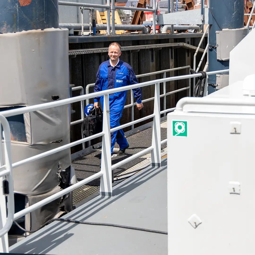 Mann in blauer Arbeitskleidung geht mit einer Tasche auf einem Schiff entlang eines Geländers.