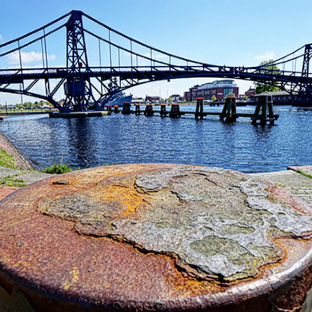 Rostige Metalloberfläche im Vordergrund mit einer blauen Brücke und Wasser im Hintergrund an einem sonnigen Tag.