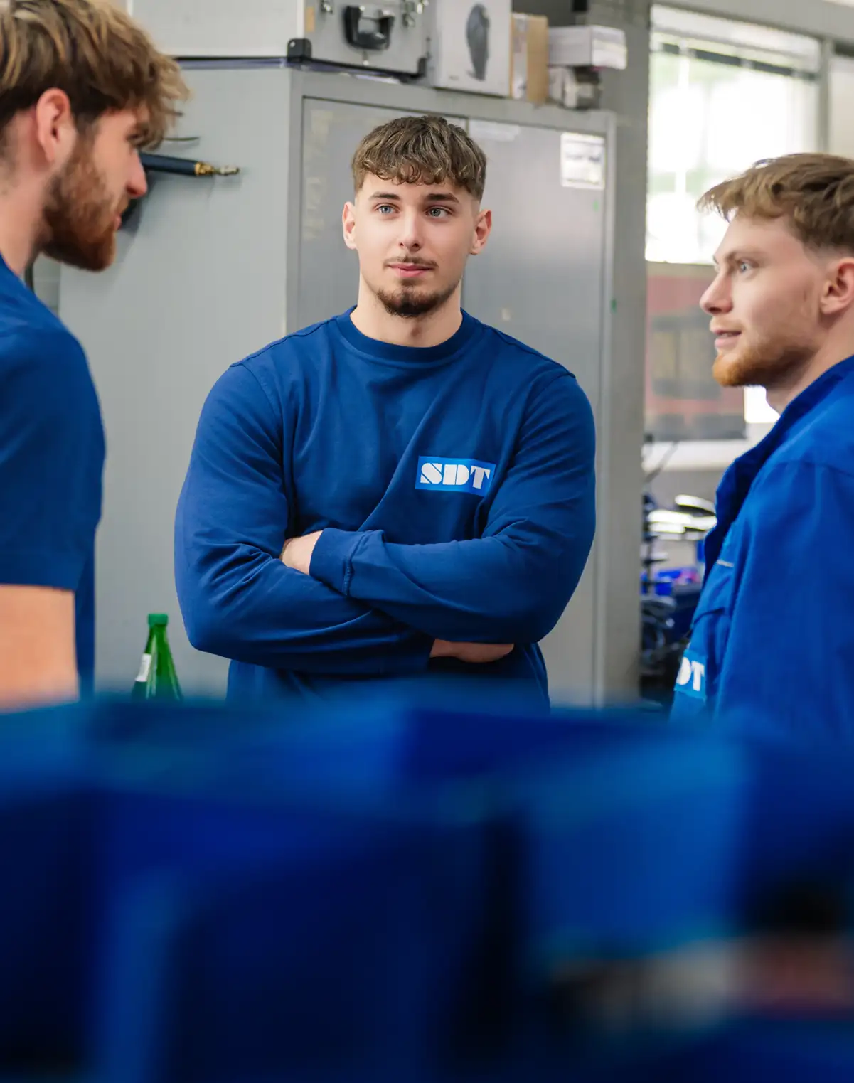 Drei Männer in blauen Arbeitskleidung unterhalten sich in einer Werkstattumgebung.