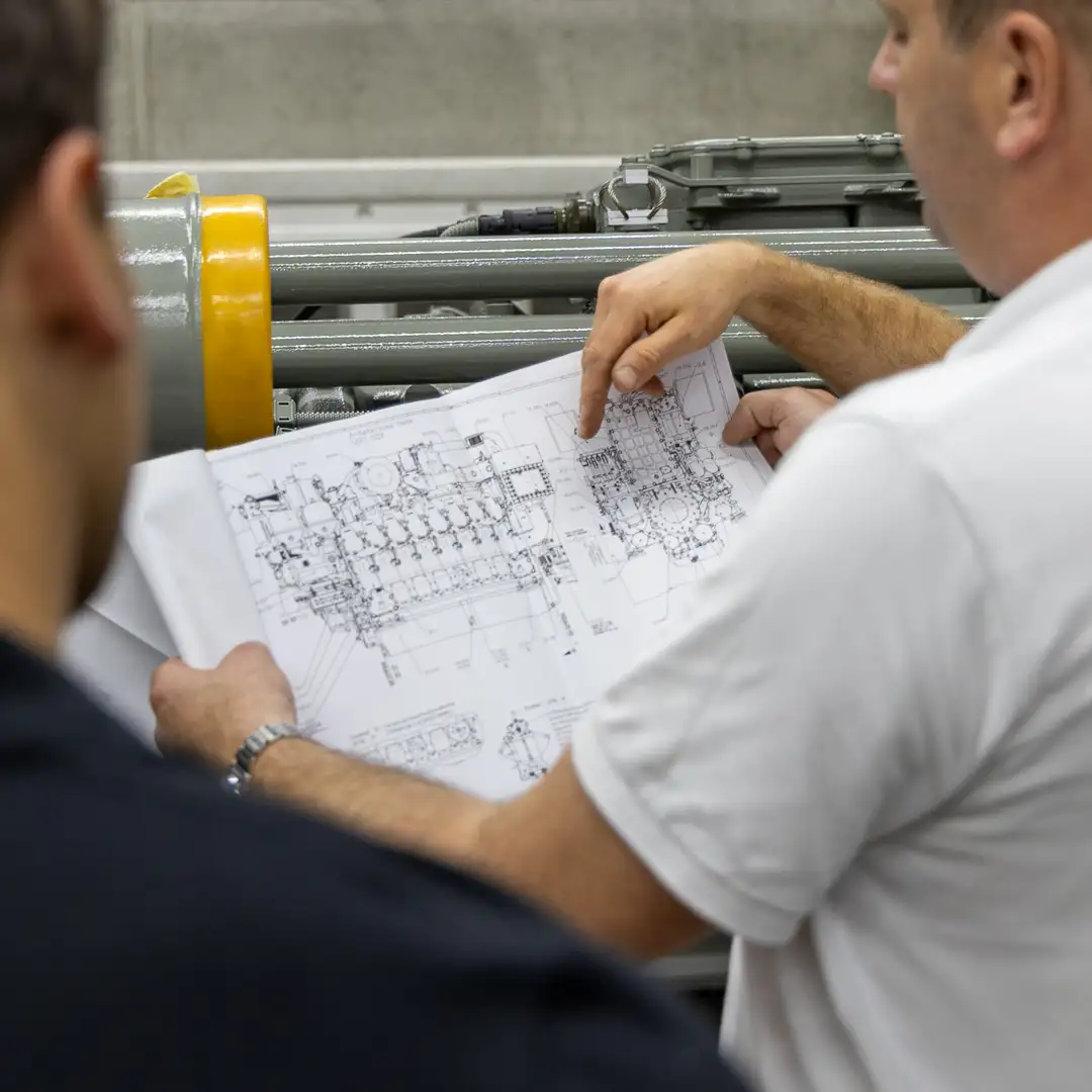 Ingenieure analysieren technische Zeichnungen vor einem großen Industriebauteil.