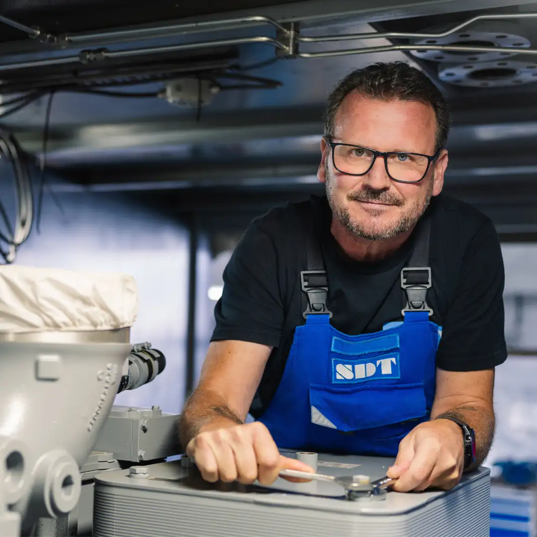Mann in blauer Arbeitskleidung arbeitet konzentriert mit Werkzeug an einer Maschine in einer industriellen Umgebung.