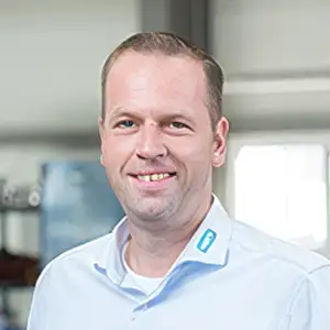 Lächelnder Mann in hellem Hemd mit Firmenlogo, stehend in einem modernen Büro.