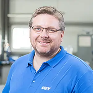 Mann in blauer Arbeitskleidung lächelt in die Kamera in einer Werkstattumgebung.