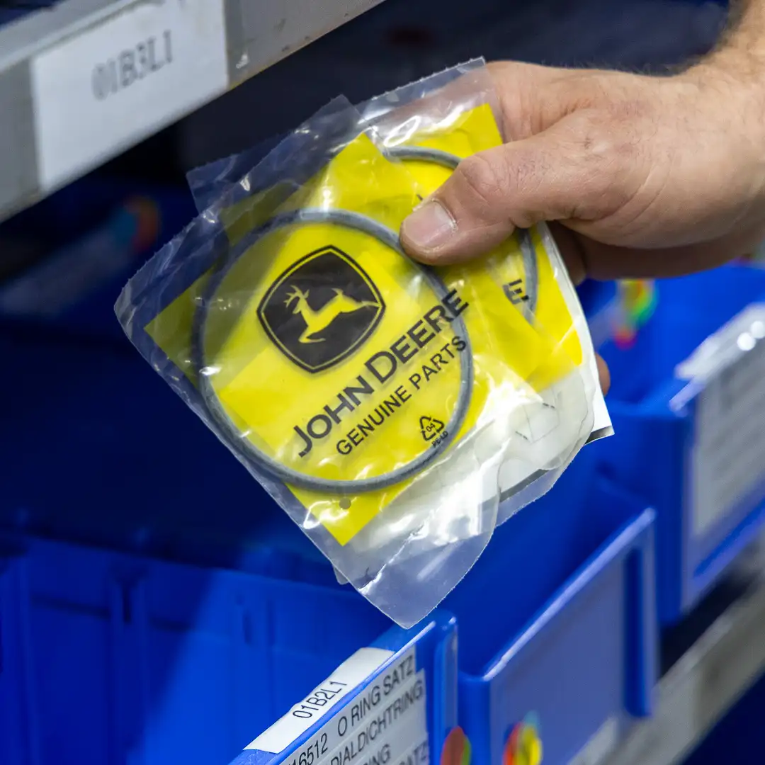 Hand hält eine John Deere Originalteile-Verpackung mit einem O-Ring vor blauen Lagerbehältern.