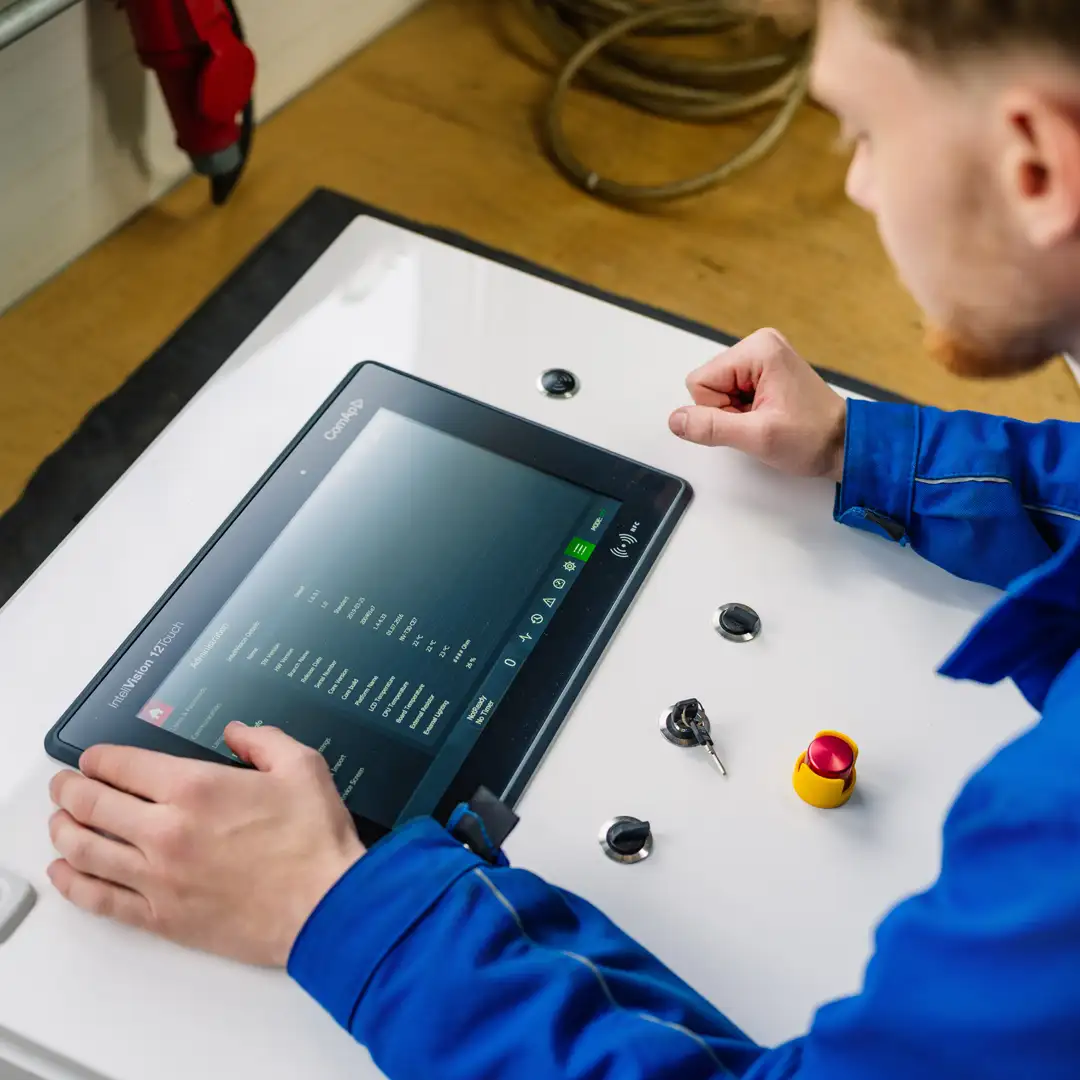 Techniker in blauer Arbeitskleidung bedient ein Kontrollpanel mit Bildschirm und Tasten in einer Werkstattumgebung.